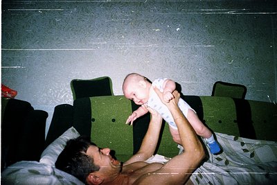 Vintage candid shot of a man lying on a bed holding an infant mid-air, 1980s-1990s. Green upholstered chairs and a patterned ...