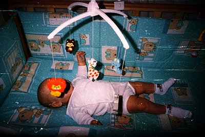 A child in a hospital crib with a mobile of cartoon animals, including a bear and a monkey, suspended above. The crib is ador...