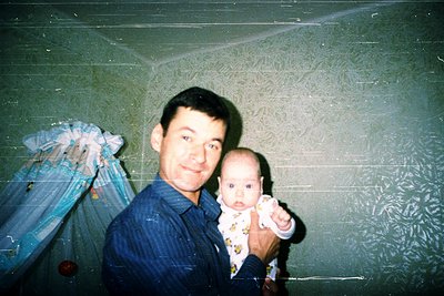 Vintage indoor portrait of a man holding a baby in a patterned onesie, likely 1980s–1990s. Textured wallpaper and a floral cu...