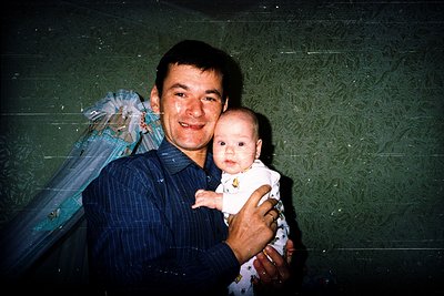 Vintage indoor portrait of a man holding a baby, likely from the 1980s–1990s. The man wears a checkered button-down shirt, an...