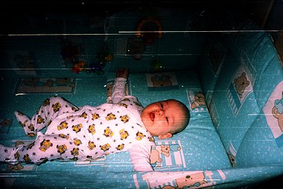 Vintage-style photo of an infant in a crib adorned with yellow-and-white cartoon bear-themed bedding, likely from the 1980s–1...