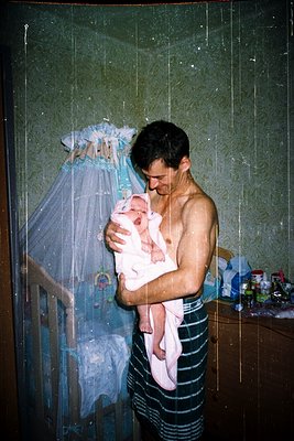 A man bathes a newborn in a modest indoor shower stall, holding the infant with a pink towel. Mosquito netting drapes over a ...