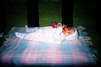 Vintage-style photo of an infant in a floral sleep sack, lying on a checkered blanket with fringe edges. The child holds a re...