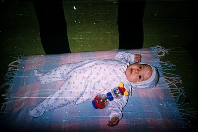 Vintage-style photo of an infant in a floral onesie, holding a toy car, lying on a fringed blanket. Warm lighting and slight ...