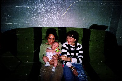Vintage candid shot of two women in a dimly lit, industrial-style interior. One holds a baby in a white outfit with a yellow ...