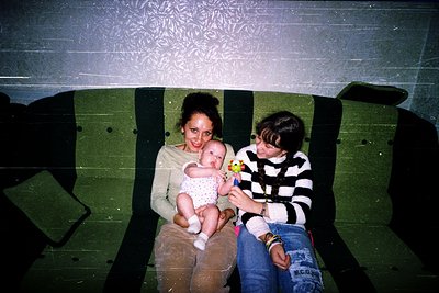 Vintage subway scene: woman cradles baby in beige sweater, man in striped sweater holds toy. Green metal seats, patterned cei...