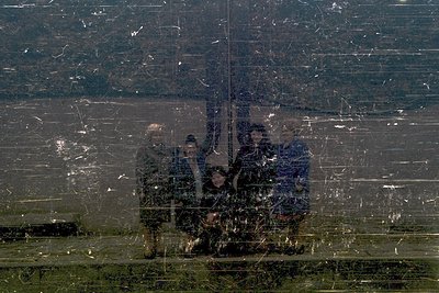 Reflection of three figures in a cracked, rain-streaked window. Urban setting with blurred buildings and greenery below. Like...