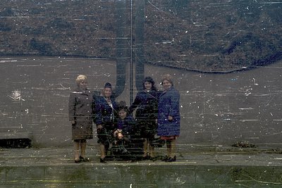 Five women pose in winter attire—coats, scarves, and boots—on a reflective glass surface, likely a monument or memorial. The ...