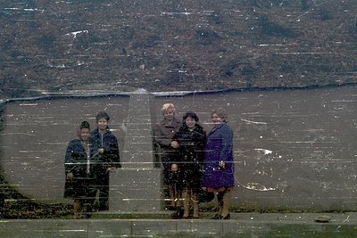 Four women in 1970s-style coats pose on a reflective surface, likely a glass wall or window, overlooking a snowy landscape. T...