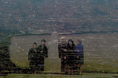 Reflection of four individuals in heavy winter coats, standing outdoors on a snowy landscape. Distinctive fur-trimmed hoods a...