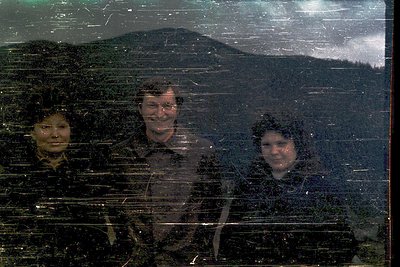 Three individuals in winter attire pose indoors, reflected in a fogged glass window showing snow-covered alpine scenery outsi...