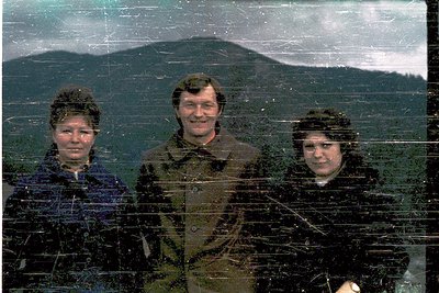 Vintage sepia-toned photo of three individuals in winter attire, posing outdoors against a snowy mountain backdrop. The centr...