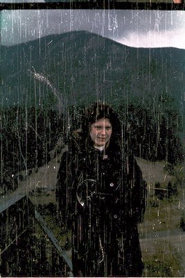 Person in heavy winter coat and hat stands in heavy rain/snowfall, smiling directly at camera. Dense forest and snow-covered ...
