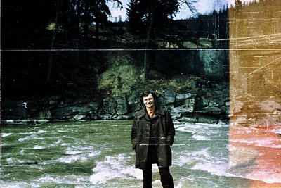 Vintage photograph of a person standing by a cascading waterfall in a forested area, framed by a rustic wooden structure. The...