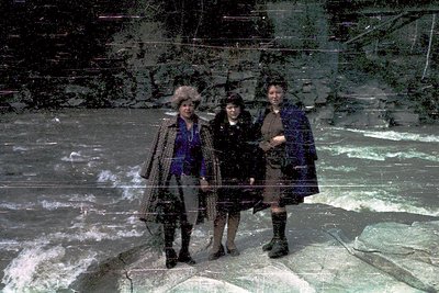 Three women pose on a frozen riverbank in winter attire, 1960s-70s. Left wears a fur hat and long coat; center in black with ...