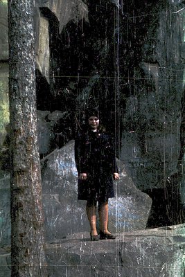 Young girl in a black dress poses on a reflective, cracked marble surface, framed by rough stone walls. The composition sugge...