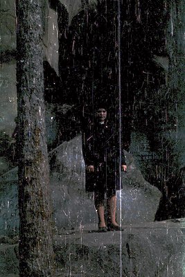 A person stands in a reflective glass corridor, holding a dark bag. The mirrored surfaces create distorted reflections of the...