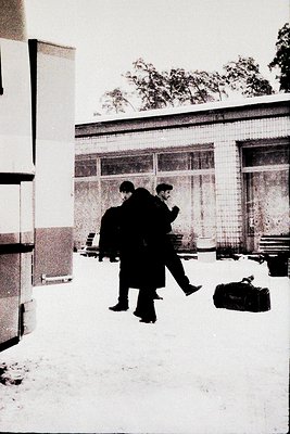 Two men in winter coats walk past a brick-façade building with large windows, carrying luggage in snowy urban setting. Mid-20...