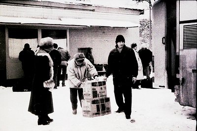 Winter street scene featuring bundled individuals carrying large cardboard boxes, likely for delivery or transport. Snow cove...
