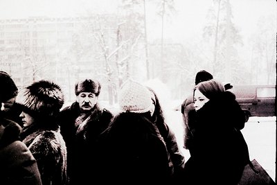 Black-and-white street scene featuring bundled individuals in winter attire—fur-lined coats, hats, and scarves—suggesting col...