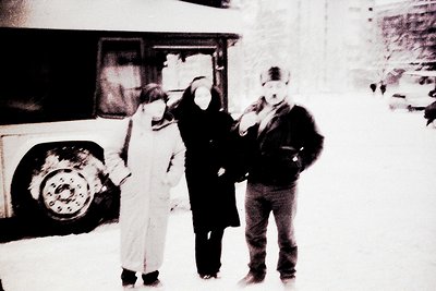 Vintage black-and-white photo of three individuals in winter attire near a bus stop, likely 1970s–1980s. Snow covers the grou...