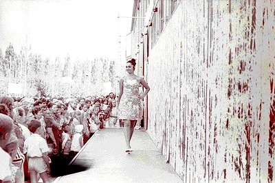 Vintage sepia-toned photo of a woman in a sleeveless, patterned dress walking a narrow pathway flanked by a crowd. The settin...
