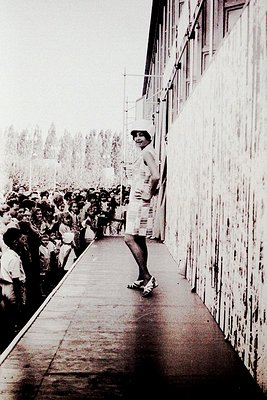 A woman in a retro striped dress and hat stands on a concrete balcony overlooking a dense crowd, likely at a public event or ...