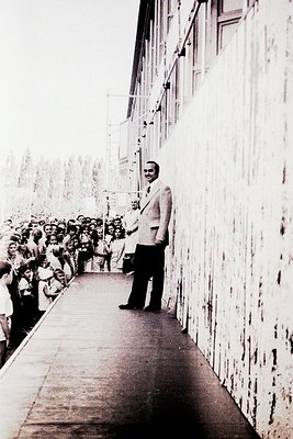 Mid-20th century black-and-white photo of a man in a light-colored suit and tie standing on a balcony overlooking a crowd. Th...