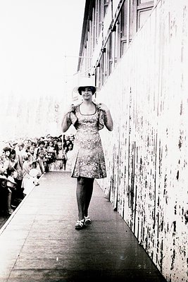 Vintage black-and-white photo of a woman in a patterned one-shoulder dress and wide-brim hat descending a ship’s gangplank, f...
