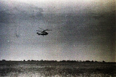 Military helicopter in mid-flight over flat, treeless terrain, likely mid-20th century. Rotor blades and tail boom visible, s...
