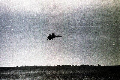 Vintage black-and-white photo of a mid-air jet, likely a fighter aircraft, in a steep climb over flat terrain. The aircraft’s...