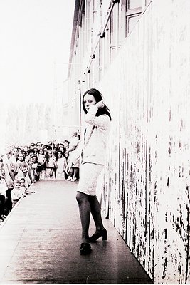 A woman in a sleeveless white dress and tights stands on a raised platform beside a weathered, textured wall, holding a micro...