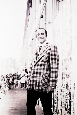 Mid-century formal portrait of a man in a plaid blazer, tie, and dark trousers, standing beside a rope barrier. Crowd of blur...