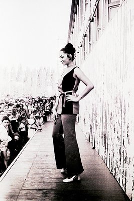 A woman in a sleek black sleeveless top and high-waisted trousers poses confidently on a narrow, elevated walkway flanked by ...