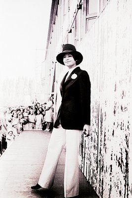 Portrait of a woman in formal attire—wide-brimmed hat, tailored blazer, and high-waisted trousers—standing on a ship’s deck. ...