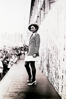 Mid-century fashion statement: woman in a striped dress and wide-brim hat strides confidently down a crowded urban corridor, ...