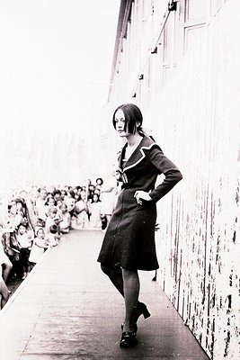 A woman in a 1960s-style sailor-inspired dress strides confidently down a crowded ship corridor, hands on hips. Crowd of pass...