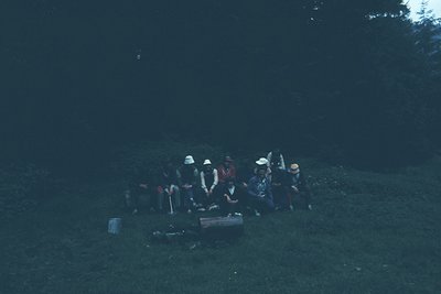 Vintage group camping scene under low light, likely evening. Eight individuals seated on grass around a fire pit with a woode...