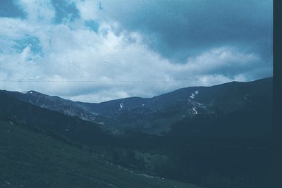 Vintage alpine landscape with rugged peaks and dense forest under dramatic, cloud-draped skies. Color saturation suggests 197...