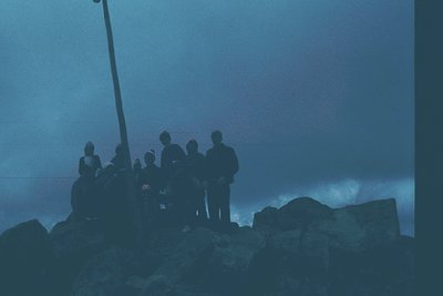Silhouetted group of 8 people standing on rocky coastline at dusk, silhouetted against misty horizon. Formal attire suggests ...