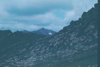 Vintage alpine landscape with rugged, snow-capped peaks under dramatic cloud cover. Foreground showcases steep, rocky terrain...