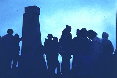 Silhouetted crowd gathered around a tall, cylindrical stone monument under a hazy blue sky. Mid-20th century attire suggests ...