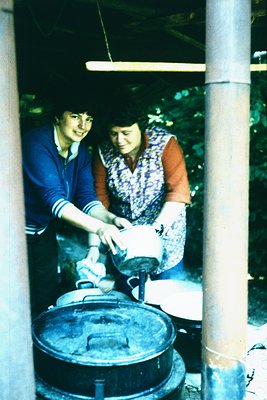 Two women in 1960s-70s outdoor kitchen, pouring liquid from a pot into a large cast-iron cauldron over an open flame. One wea...