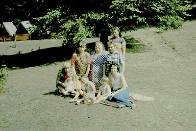 Vintage family portrait in outdoor setting, likely 1960s-1970s. Six individuals pose on sandy ground near rustic wooden cabin...