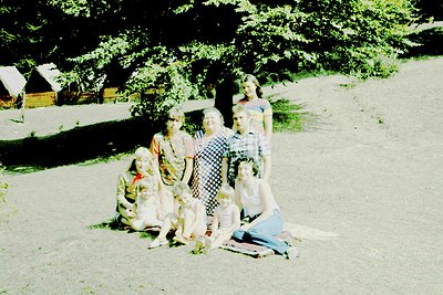 Vintage group portrait in outdoor setting, likely 1960s–1970s. Five individuals pose on a blanket, surrounded by rustic woode...