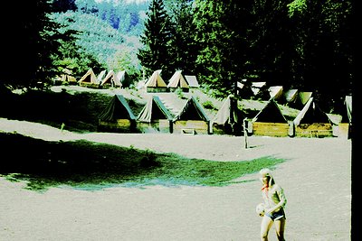 Vintage alpine camping site with canvas tents arranged in a grid, surrounded by dense forest. A lone woman in mid-stride, wea...