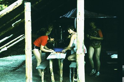 Vintage outdoor scene featuring three individuals playing a board game under a makeshift canopy. Wooden structure with rustic...
