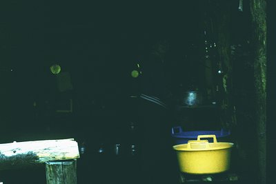 Low-light interior shot of a rustic wooden structure, likely a barn or storage shed. A yellow bucket and blue metal container...
