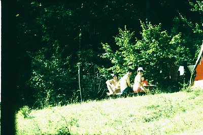 Vintage outdoor scene featuring three individuals seated on grassy hillside under dense trees. One wears a white hat, another...
