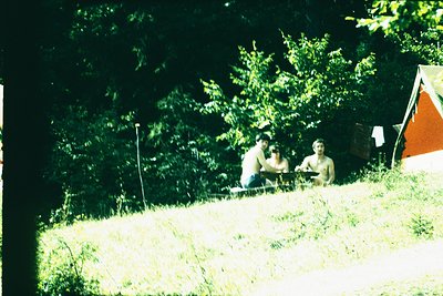 Vintage camping scene with three individuals seated on grassy hillside, surrounded by dense forest. A vintage-style tent in b...